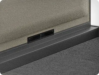 Ouverture de ventilation au niveau du sol intégrée à la base d’une cabine de bureau, présentée en détail avec cadre foncé et sol textile.